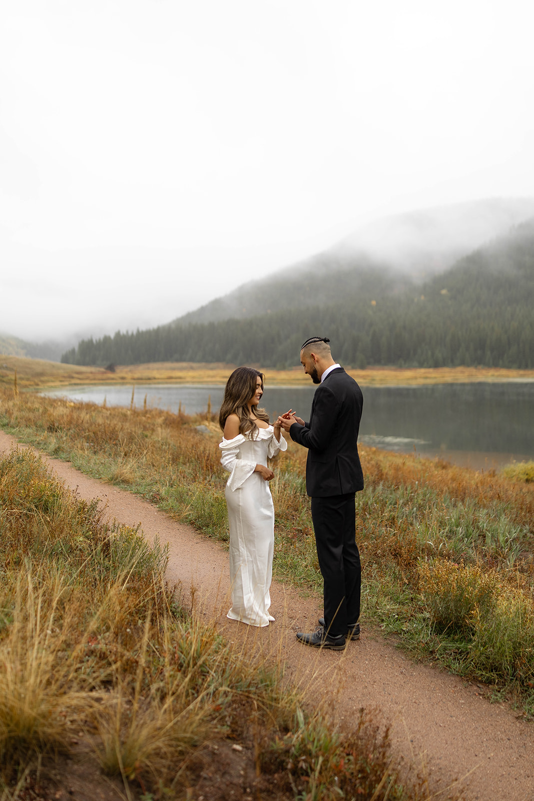 A bride and groom share a quiet moment exchanging rings on the lakeside trail at Piney River Ranch, framed by autumn colors and low clouds.