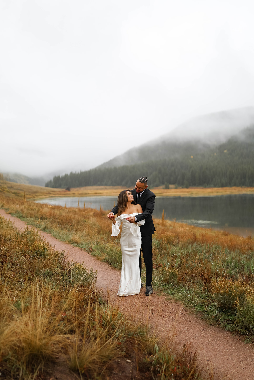 A bride and groom walking hand in hand along the misty shoreline at Piney River Ranch with pine-covered mountains behind them.