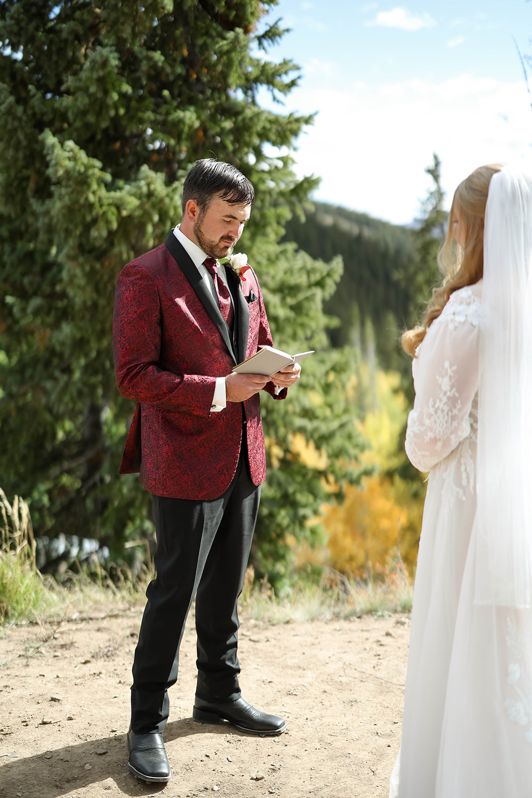Groom reading his vows from a small notebook as the bride listens, surrounded by evergreens at their aspen elopement ceremony.