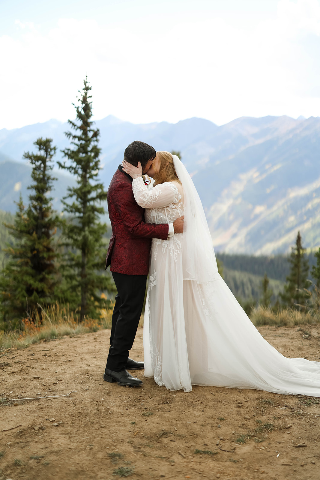 Bride and groom sharing a quiet kiss on a dirt overlook with sweeping Colorado mountain views behind them.