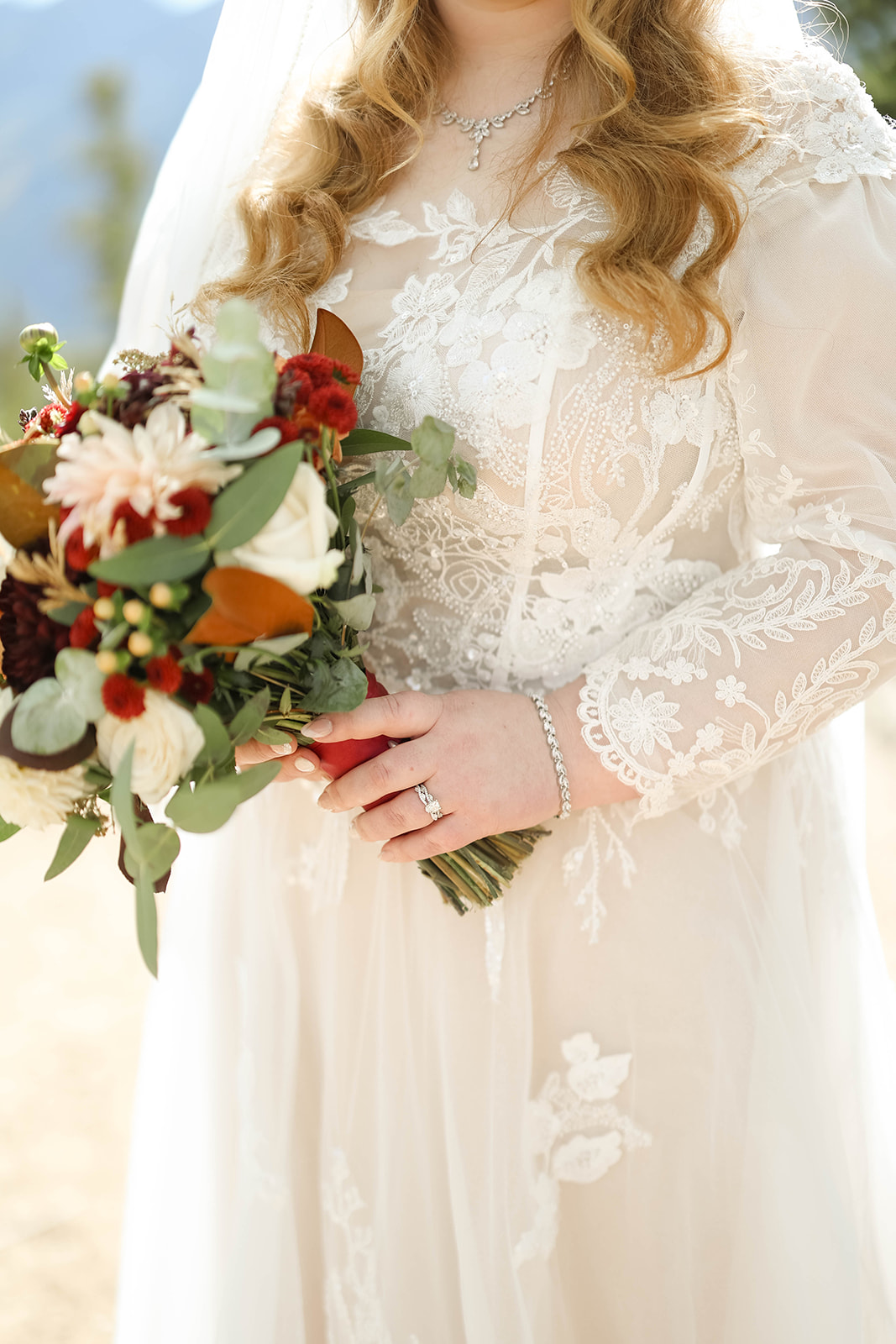 Close-up of a bride holding a fall-inspired bouquet, highlighting lace sleeve details, engagement ring, and soft floral textures.
