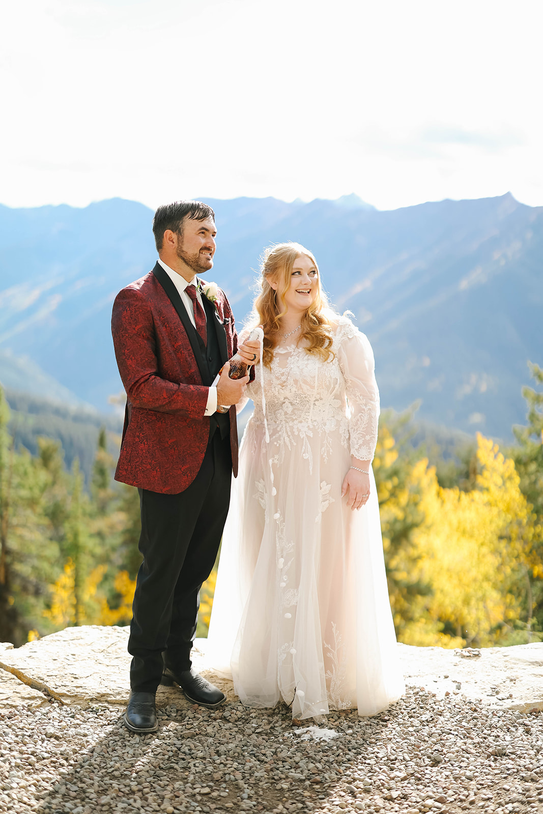 Bride and groom standing on a mountain overlook during their aspen elopement, holding a champagne bottle with sweeping alpine views and golden fall foliage behind them.