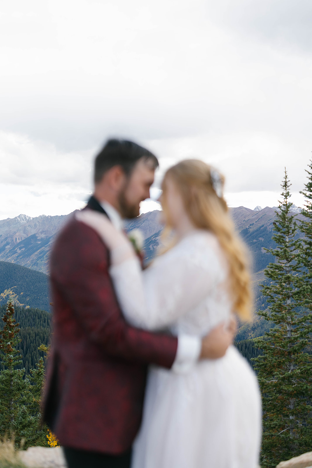 Soft, intimate portrait of a newly married couple embracing on a mountain ridge, framed by evergreen trees and layered blue peaks in the distance.