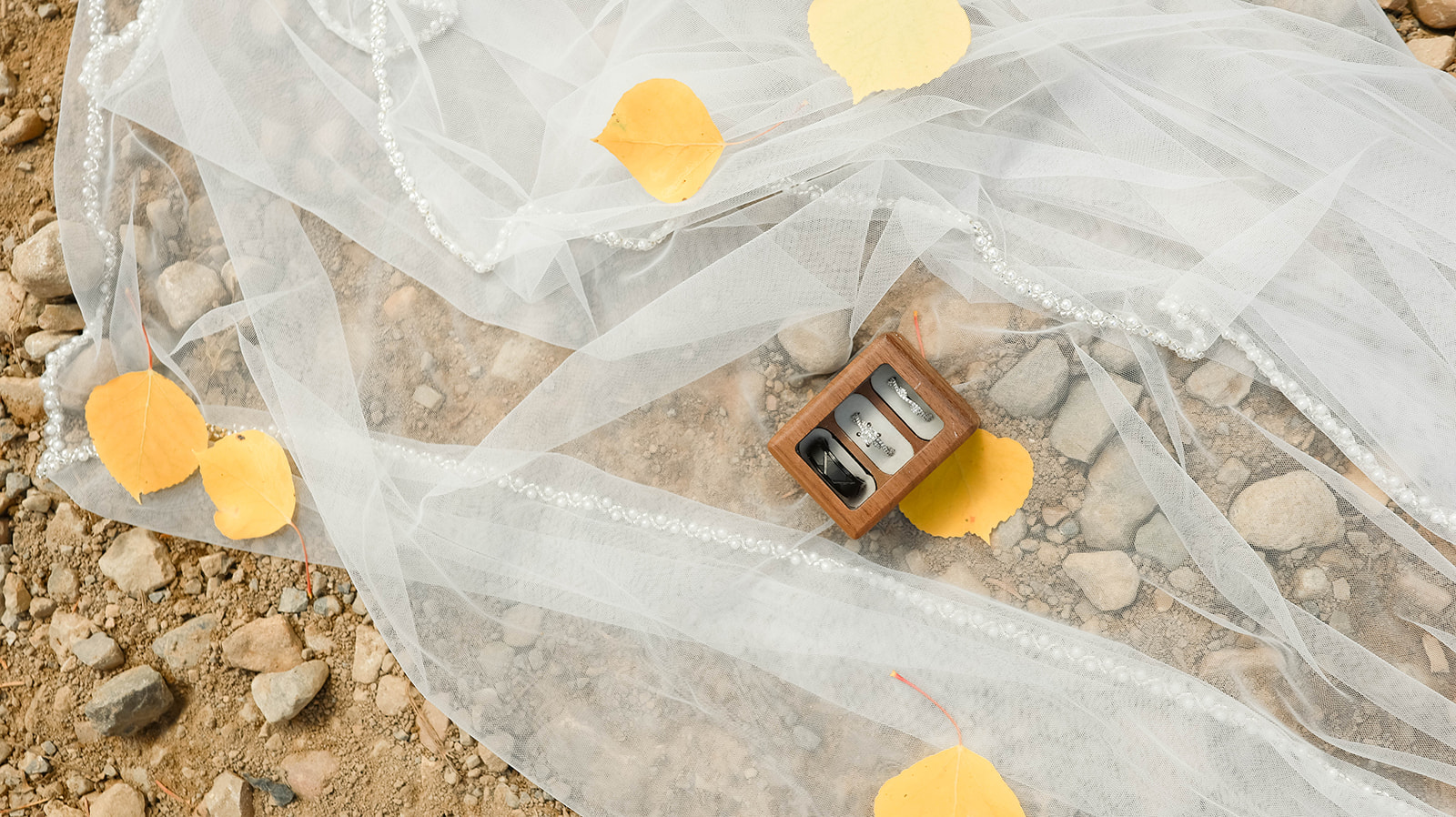 Flat lay of wedding rings resting on a sheer veil with fall leaves scattered across rocky ground.