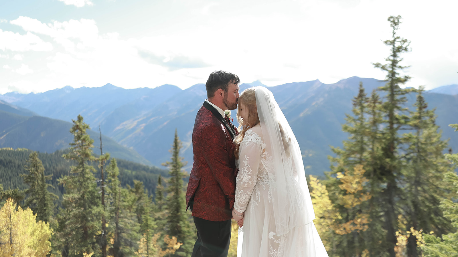 Groom kissing the bride’s forehead as they hold hands on a scenic Colorado mountaintop, surrounded by tall pines and early autumn color.