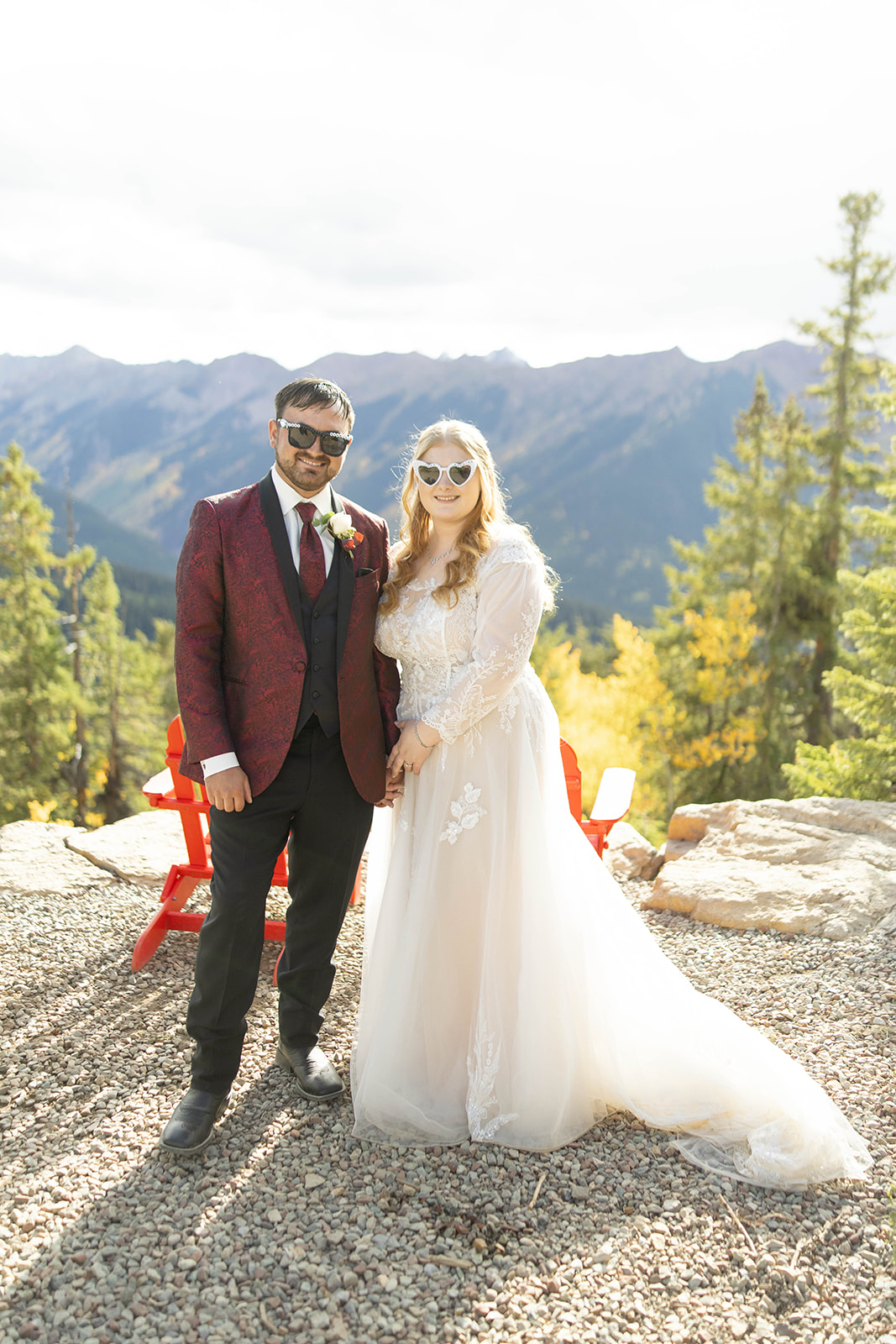 Newlyweds standing together in sunglasses and wedding attire on a gravel overlook, smiling with mountains behind them at their aspen elopement.