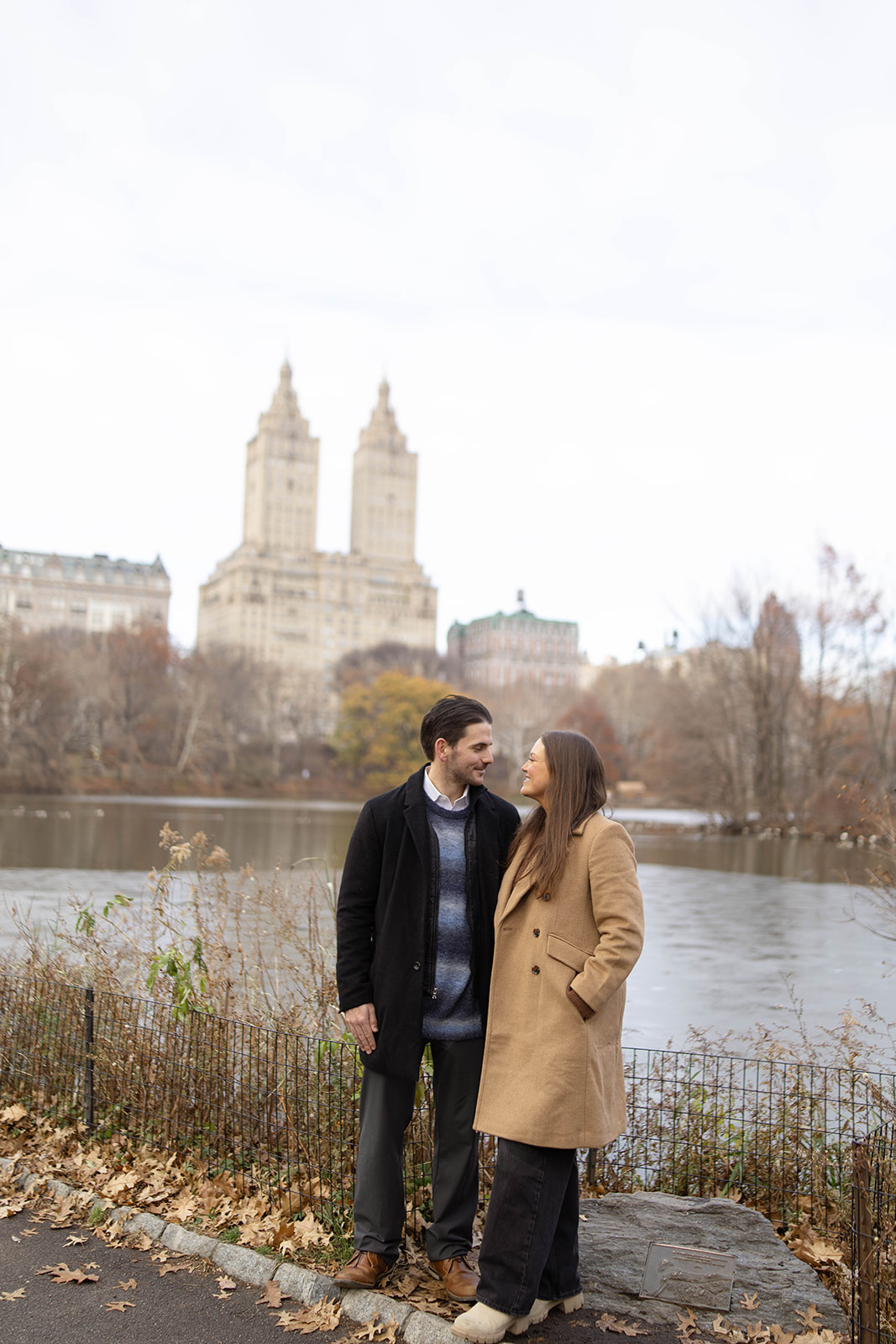 Couple standing together by the water, smiling at each other with iconic Upper West Side buildings in the background.