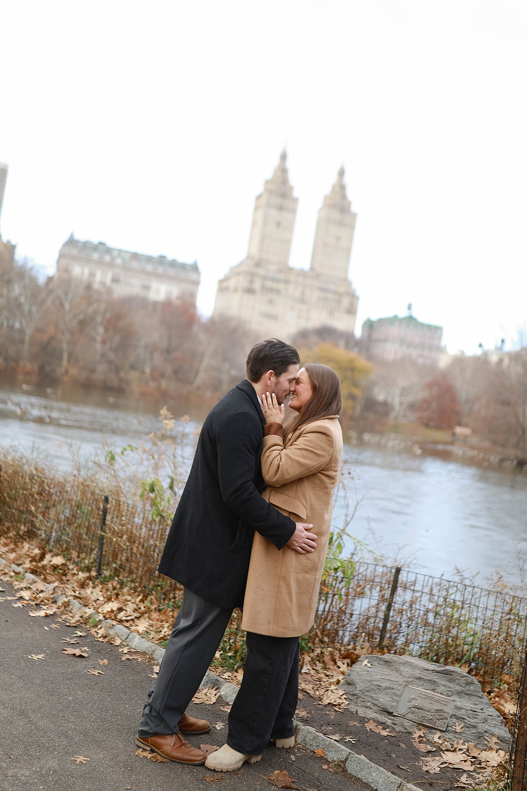 Couple standing close along the lake path in Central Park, framed by fall leaves and historic buildings in the background.