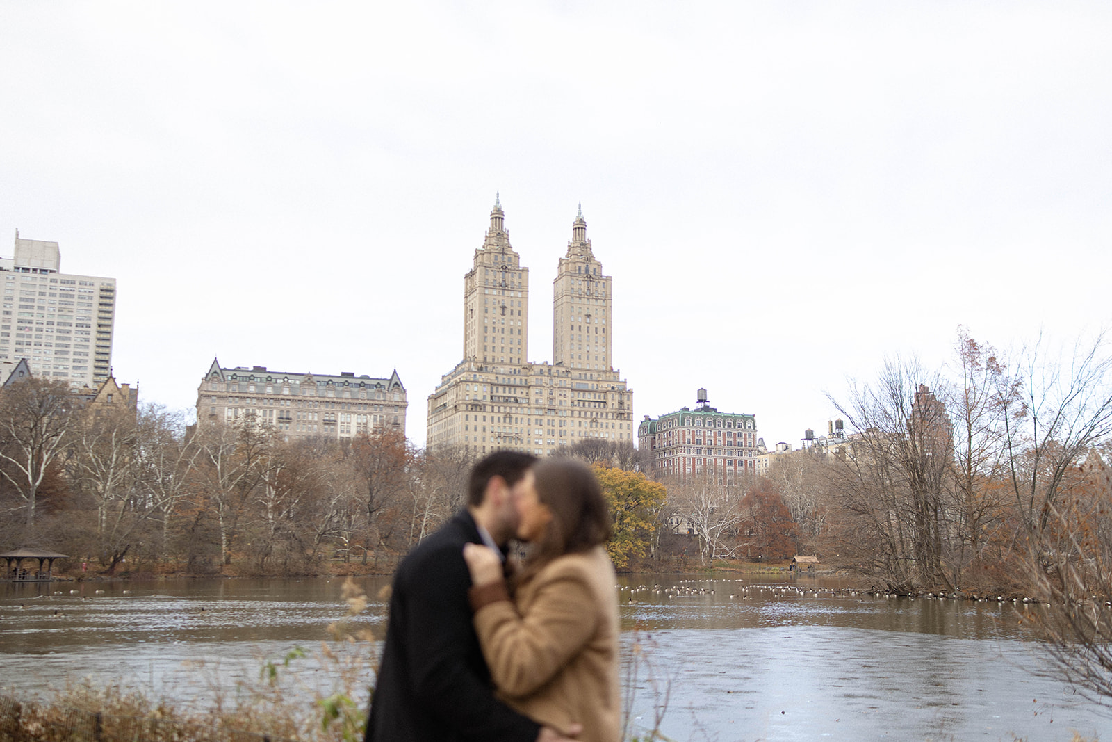 A wide shot of the couple kissing by the water with classic Upper West Side buildings rising behind them during a romantic central park engagement.