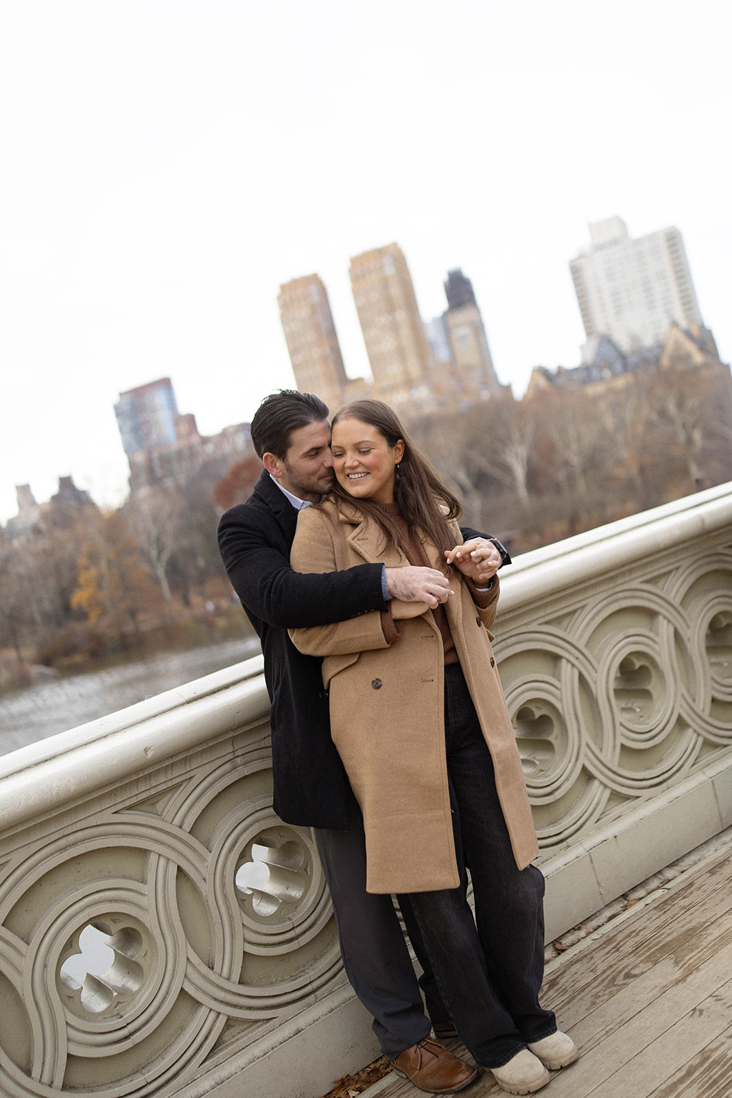 A cozy embrace on a decorative bridge as the city skyline peeks through bare winter trees during a central park engagement shoot.