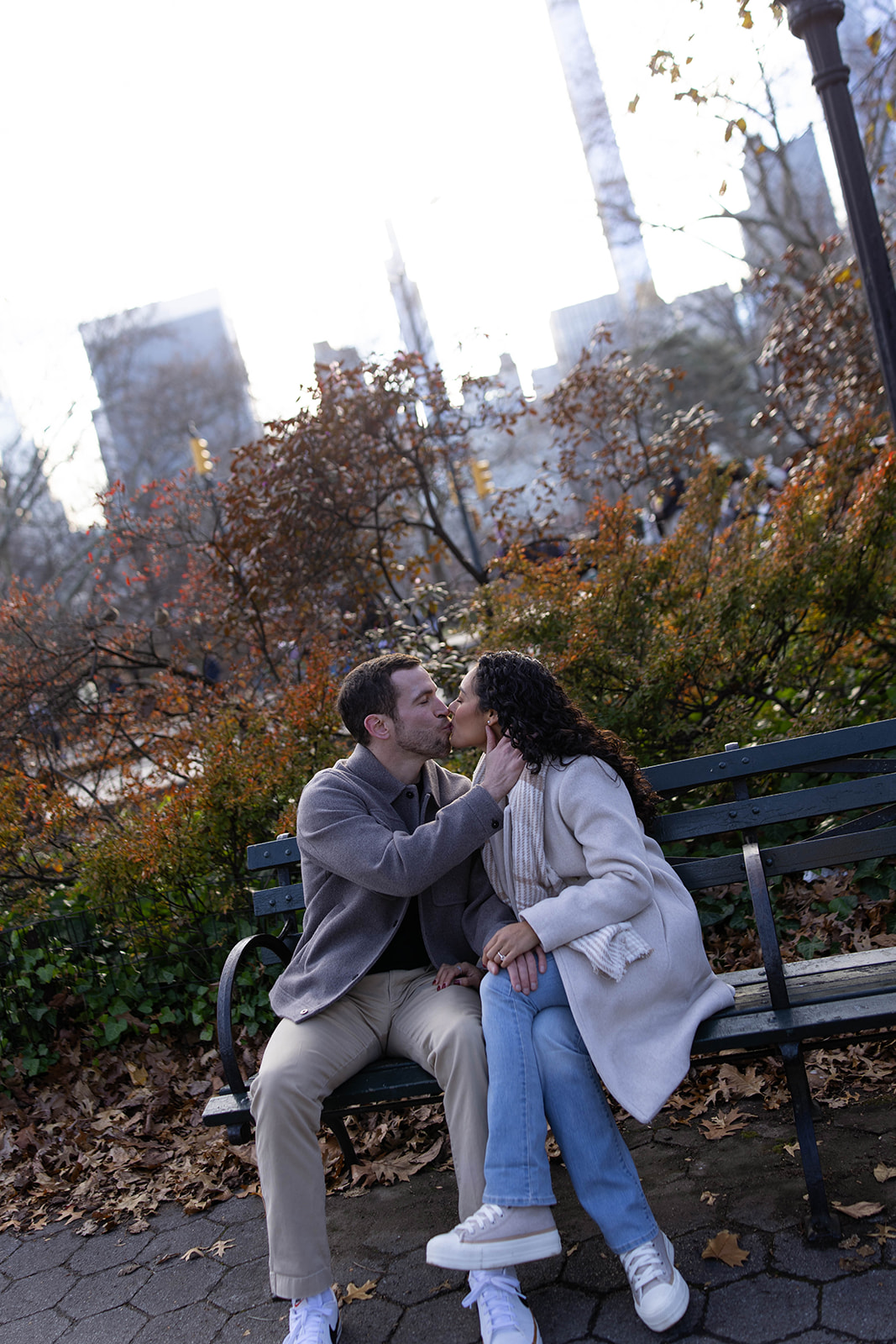 Couple sharing a kiss on a park bench surrounded by fall foliage during a cozy central park engagement.
