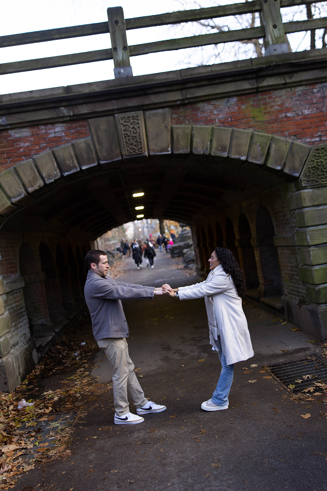 Playful moment of a couple holding hands beneath a stone bridge during their central park engagement photos.