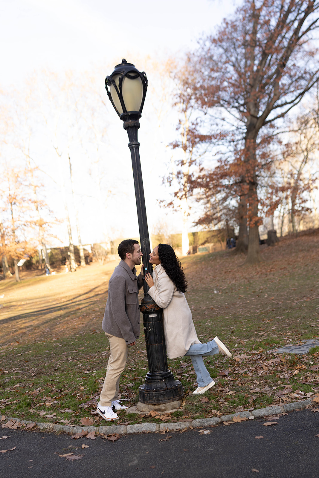 Playful engagement photo of a couple posing by a vintage lamppost in Central Park, with fallen leaves and autumn tones around them.