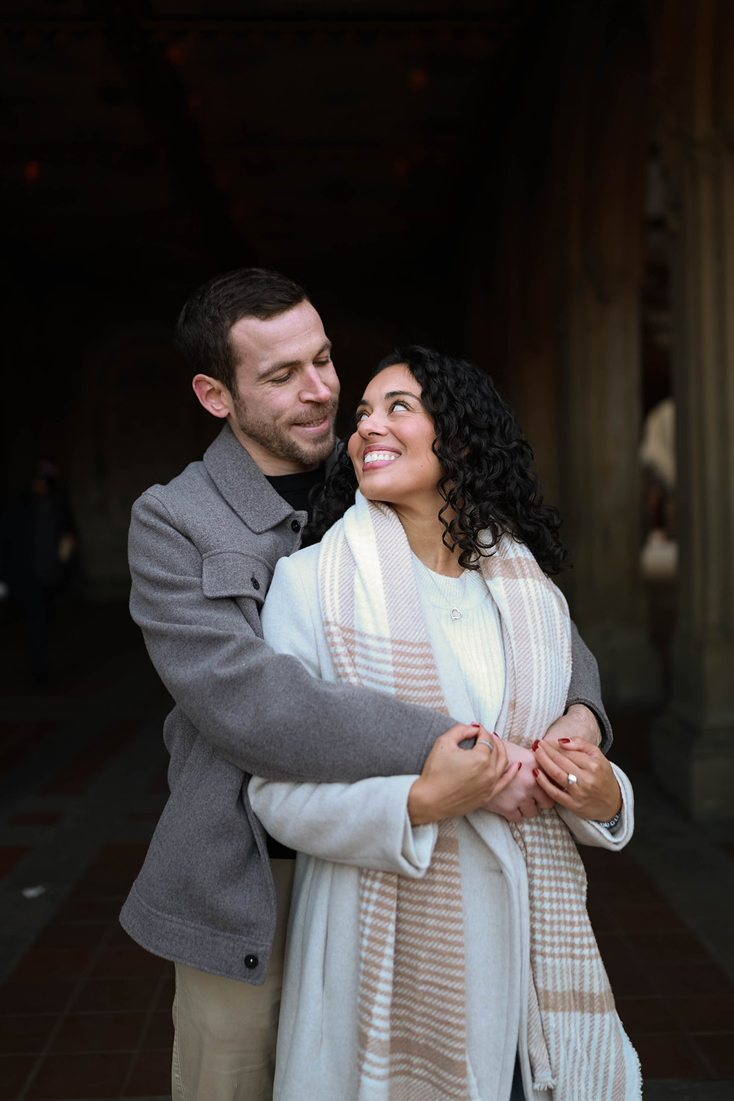 Couple wrapped in a warm embrace under an arched walkway, smiling softly at each other in a quiet, intimate moment.
