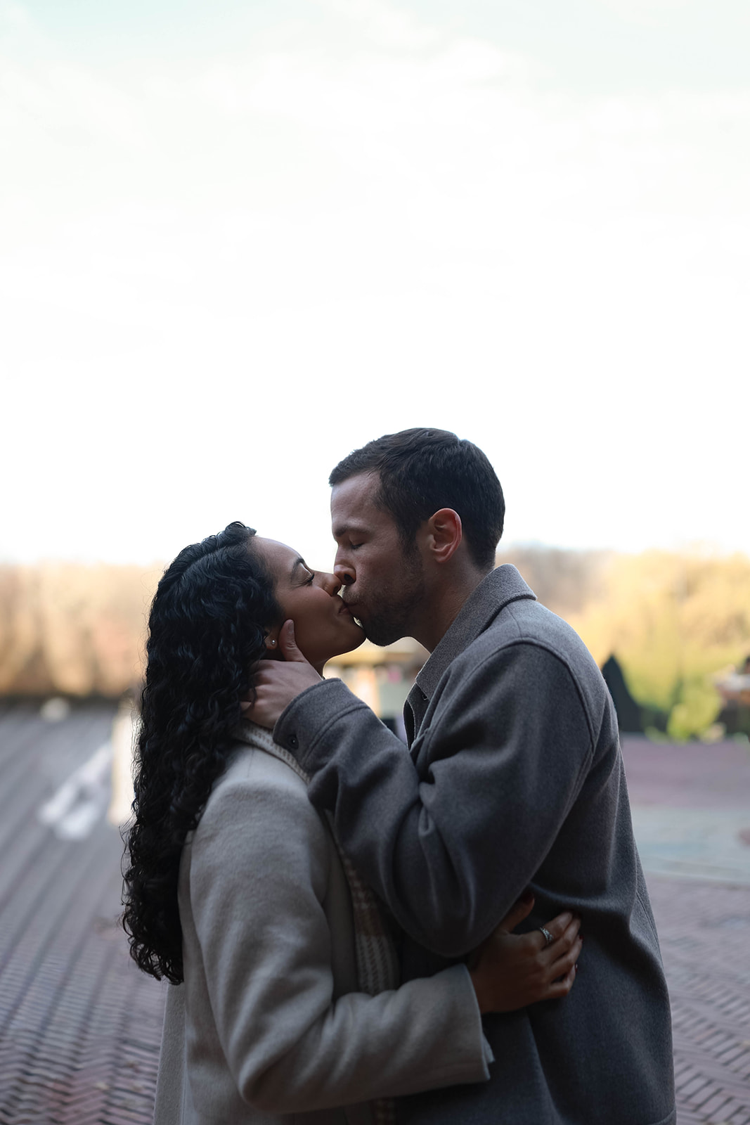 Couple sharing a quiet kiss on a brick terrace during their central park engagement, wrapped in soft winter layers with the city blurred behind them.