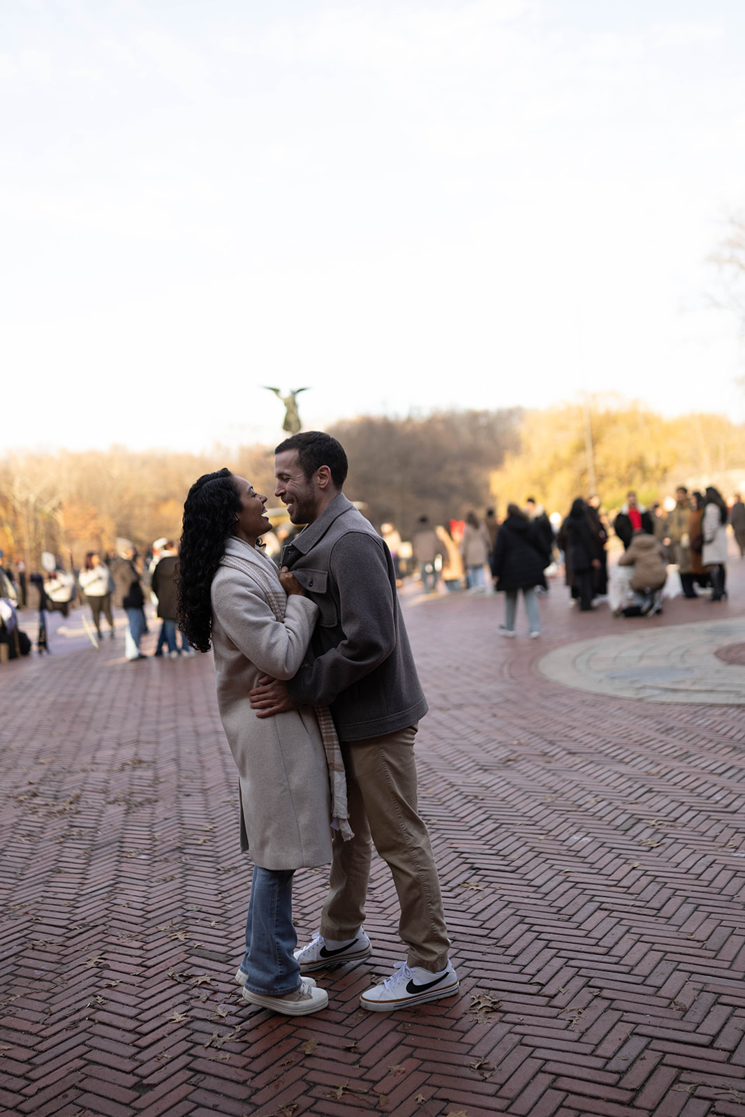 A couple standing close and laughing together at Bethesda Terrace, surrounded by the iconic brick plaza and winter trees.