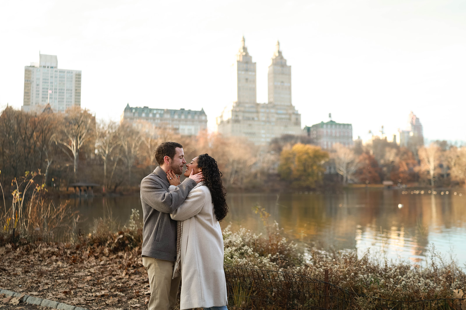 Intimate embrace by the water with the Upper West Side skyline reflected behind them, highlighting a peaceful engagement moment.