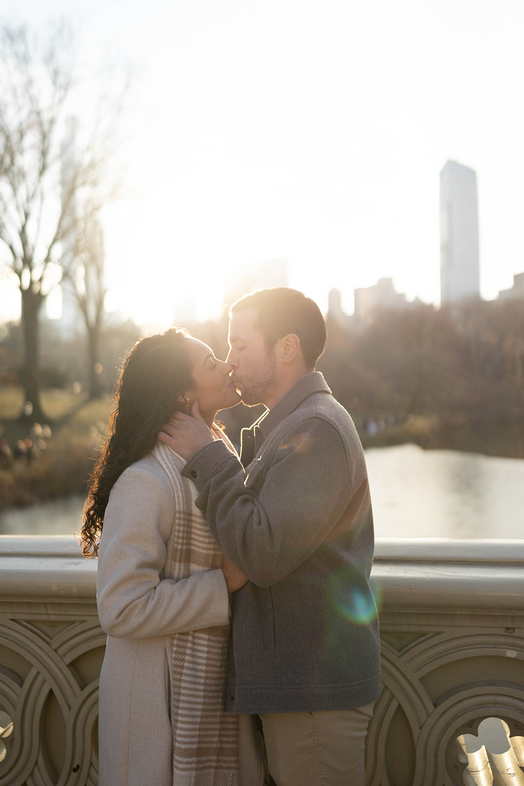 The couple shares a sunlit kiss by the water, with golden light flaring behind them and the city softly blurred in the distance.