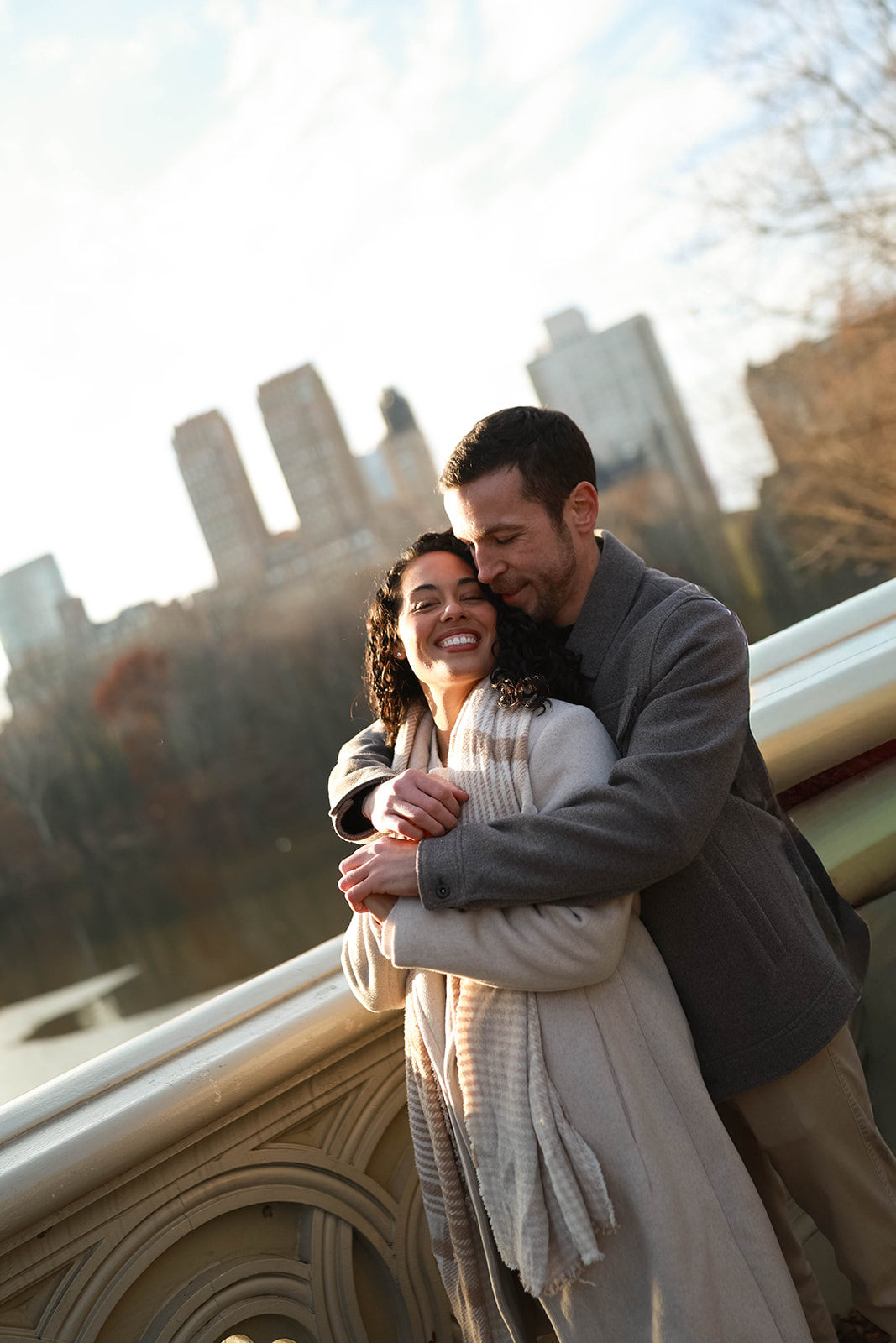 A couple cuddles on a stone bridge overlooking the water with the New York City skyline glowing behind them during a central park engagement session.
