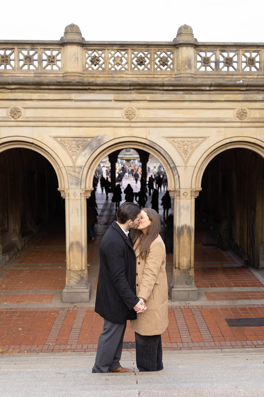 The couple shares a kiss beneath the iconic Bethesda Terrace arches, framed by warm stone details and passersby in the background.