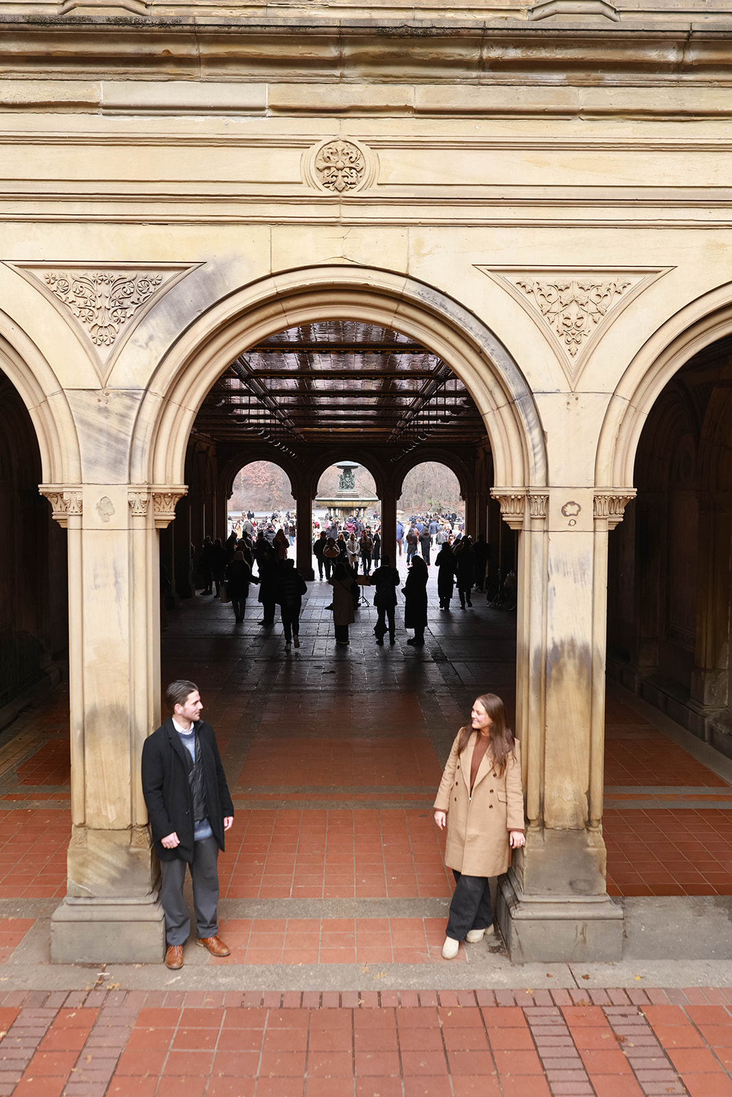 The couple stands beneath the Bethesda Terrace arches, smiling at each other across the walkway during a timeless central park engagement session.