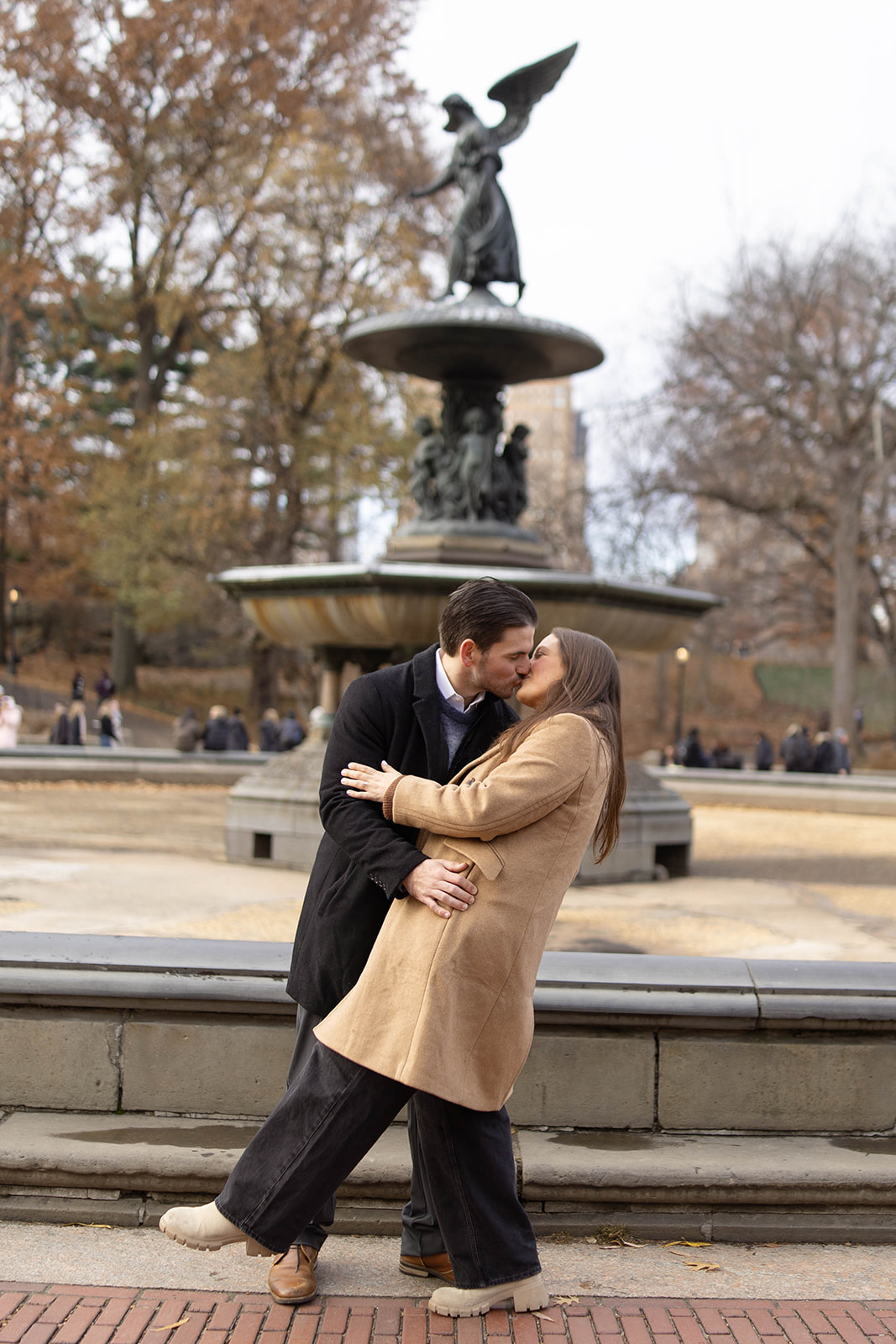 Couple kissing in front of Bethesda Fountain during a central park engagement, surrounded by winter trees and classic stone details.