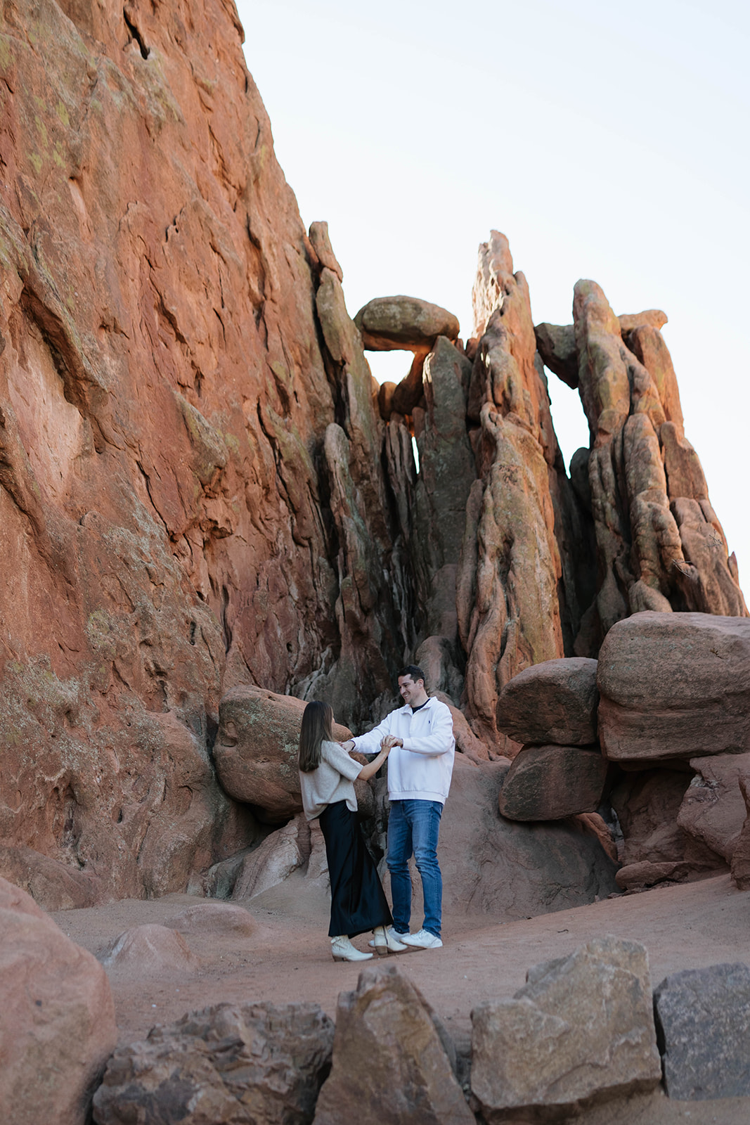 A playful scene of a couple holding hands and dancing between towering sandstone walls, surrounded by the unique rock textures of Garden of the Gods.