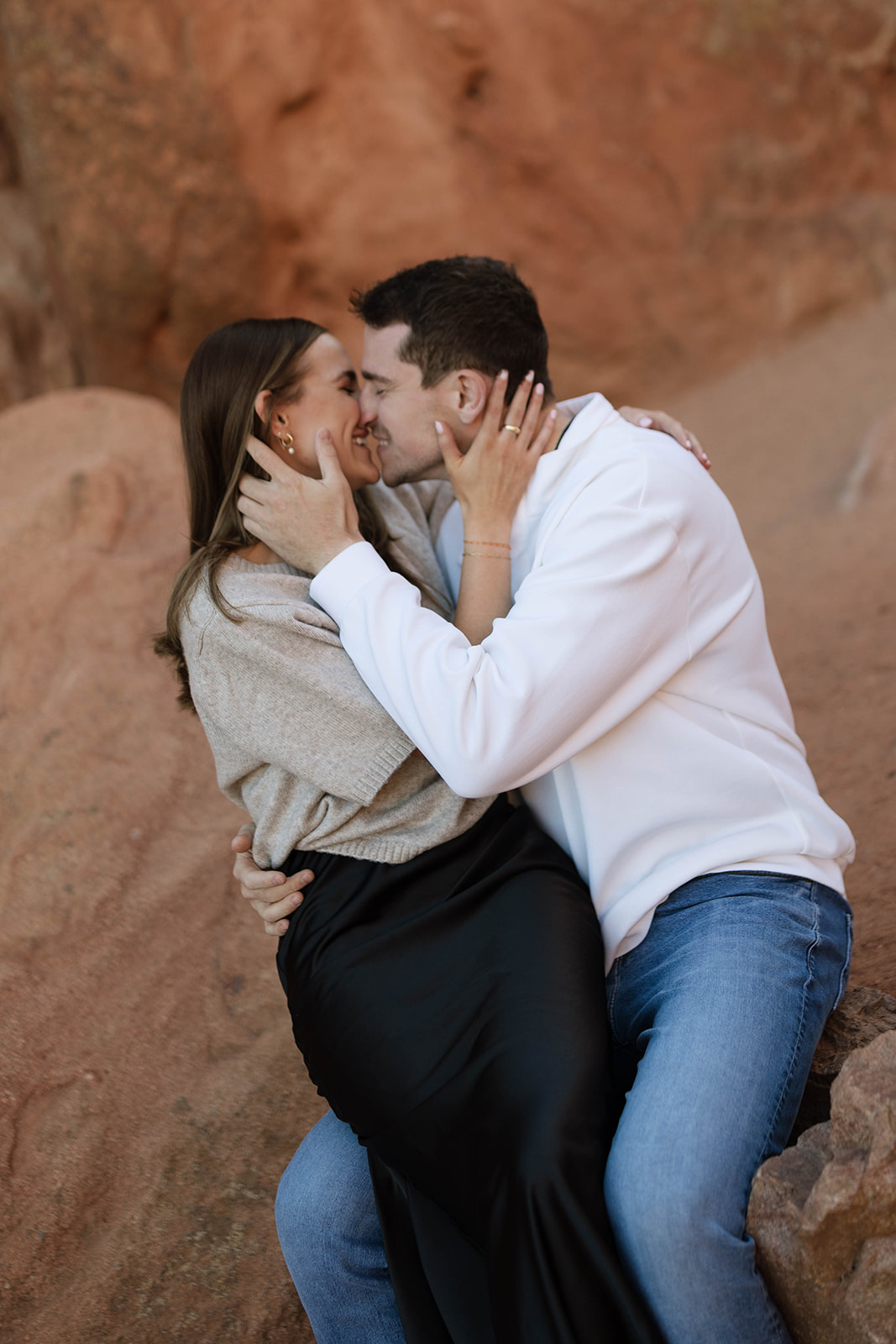A couple sharing a close, joyful kiss while sitting together on warm red rocks, holding each other with big smiles.