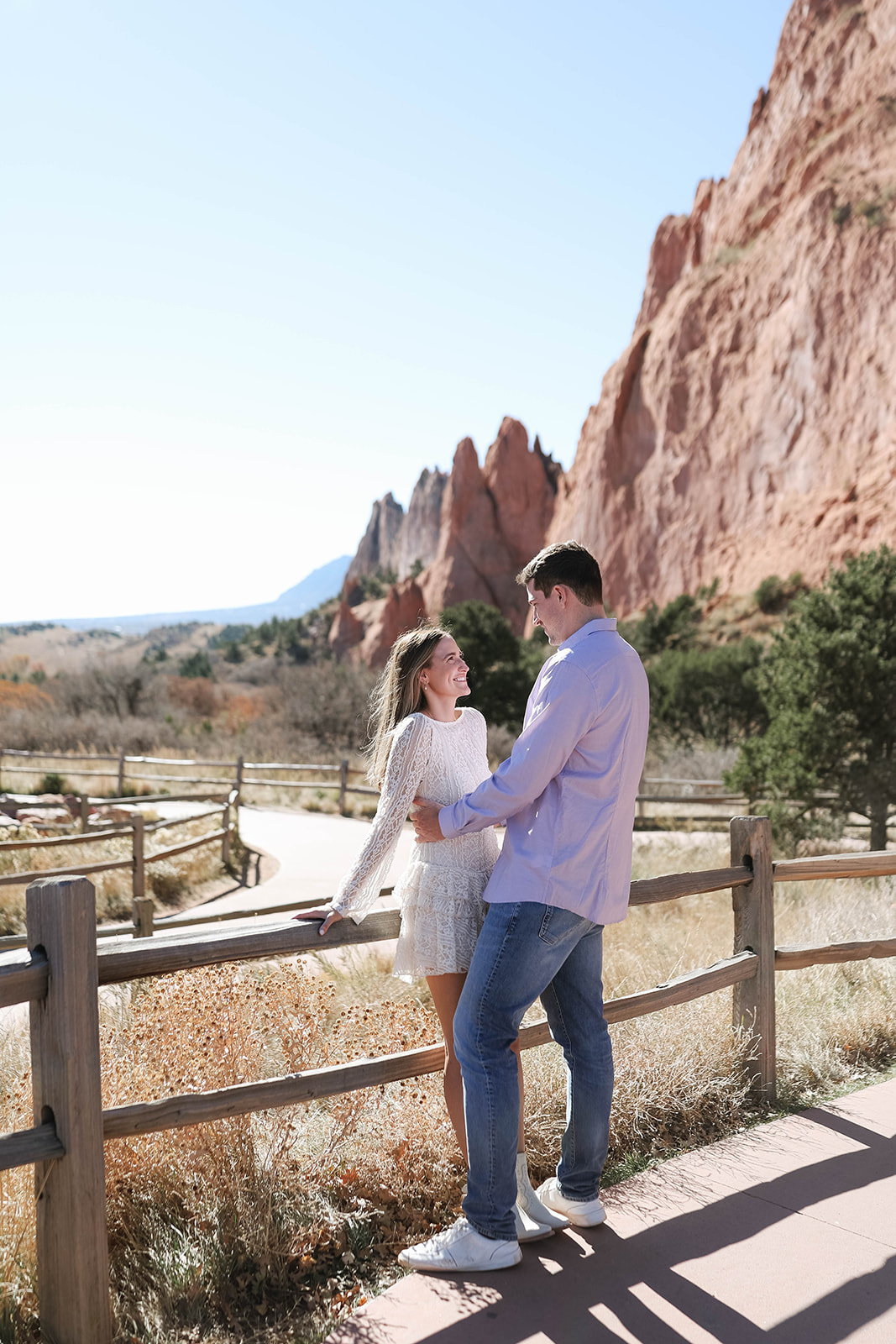 A joyful couple smiling at each other near a wooden fence with the breathtaking red cliffs of Garden of the Gods behind them.