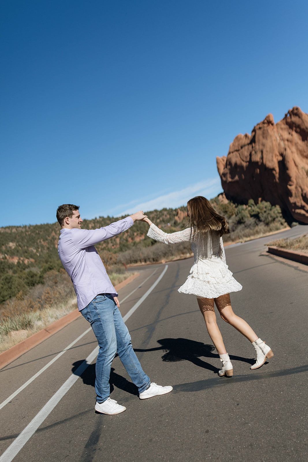 A playful moment of a couple spinning each other in the middle of a sunlit road with the red rock formations of Garden of the Gods rising behind them.