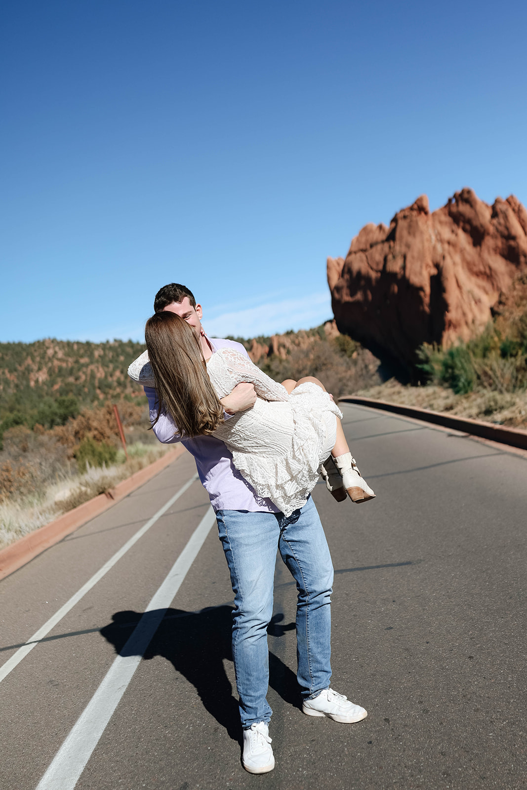 A man lifting his partner into his arms on a sunlit road bordered by iconic red rock formations in Garden of the Gods.