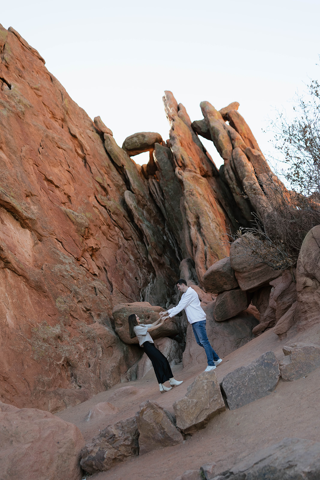 A couple exploring a rugged rock formation together, playfully reaching for each other on a sloped sandstone ledge at Garden of the Gods.