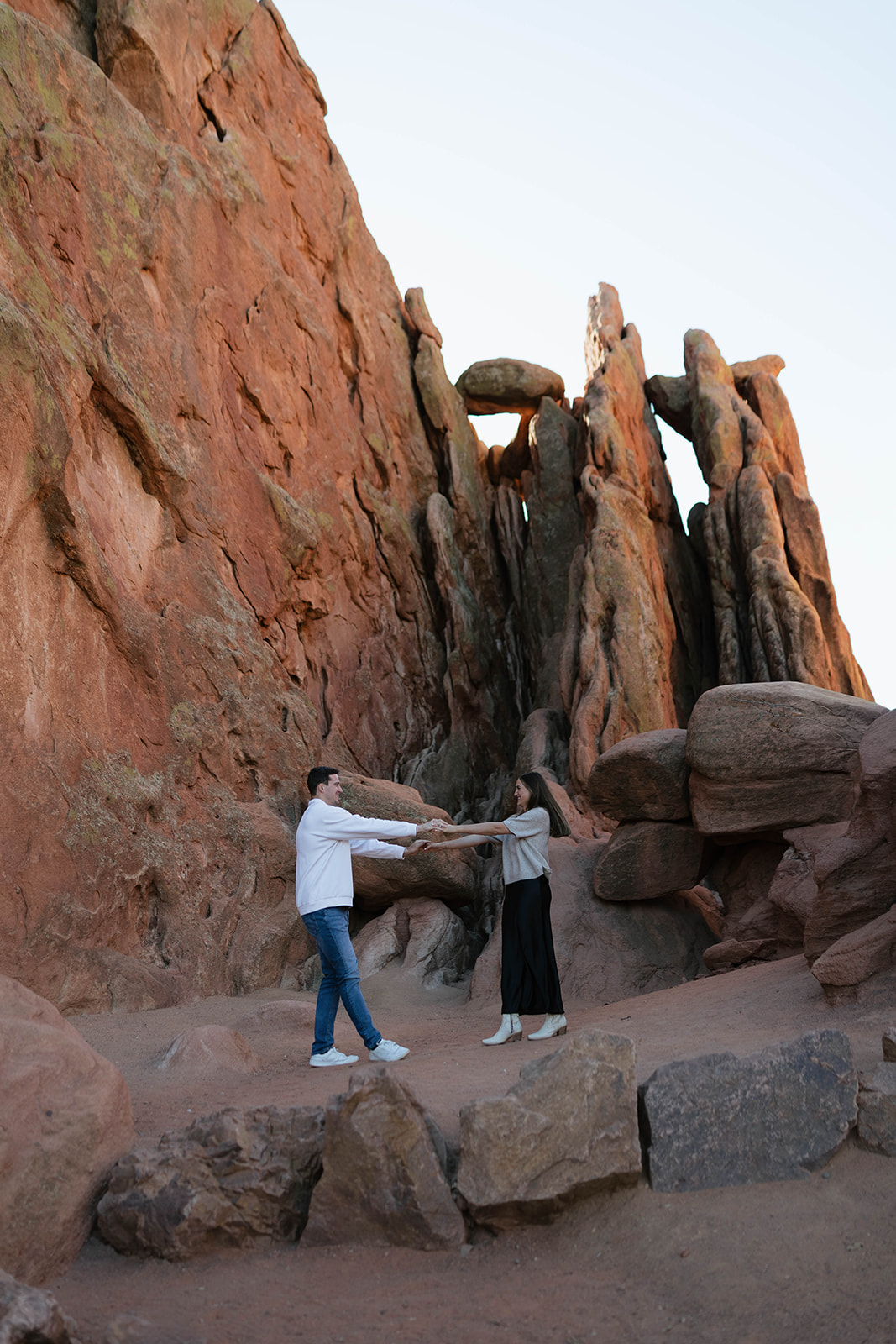 A couple holding hands and playfully leaning toward each other among massive, rugged red rock formations.