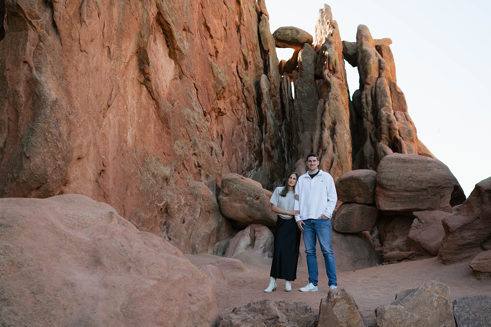 A couple standing together at the base of massive red rock walls, framed by rugged textures and soft natural light.