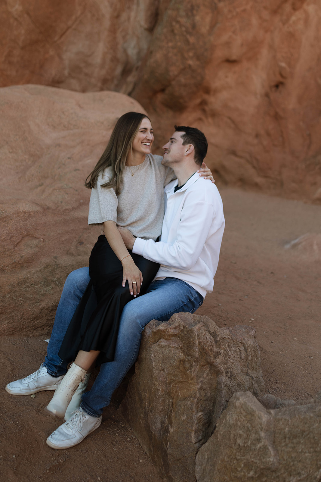 A couple sitting together on warm red rocks, smiling and leaning in close as they enjoy the peaceful desert setting.