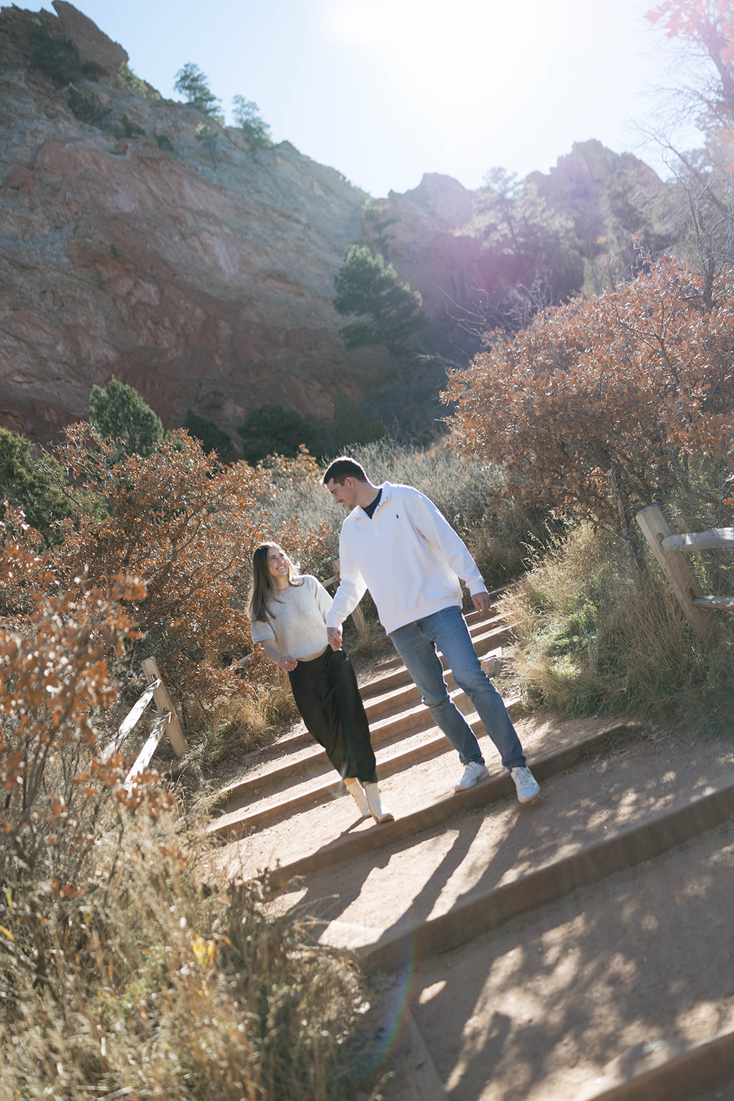 A couple holding hands and walking down sunlit stone steps surrounded by golden fall foliage and towering red rocks.