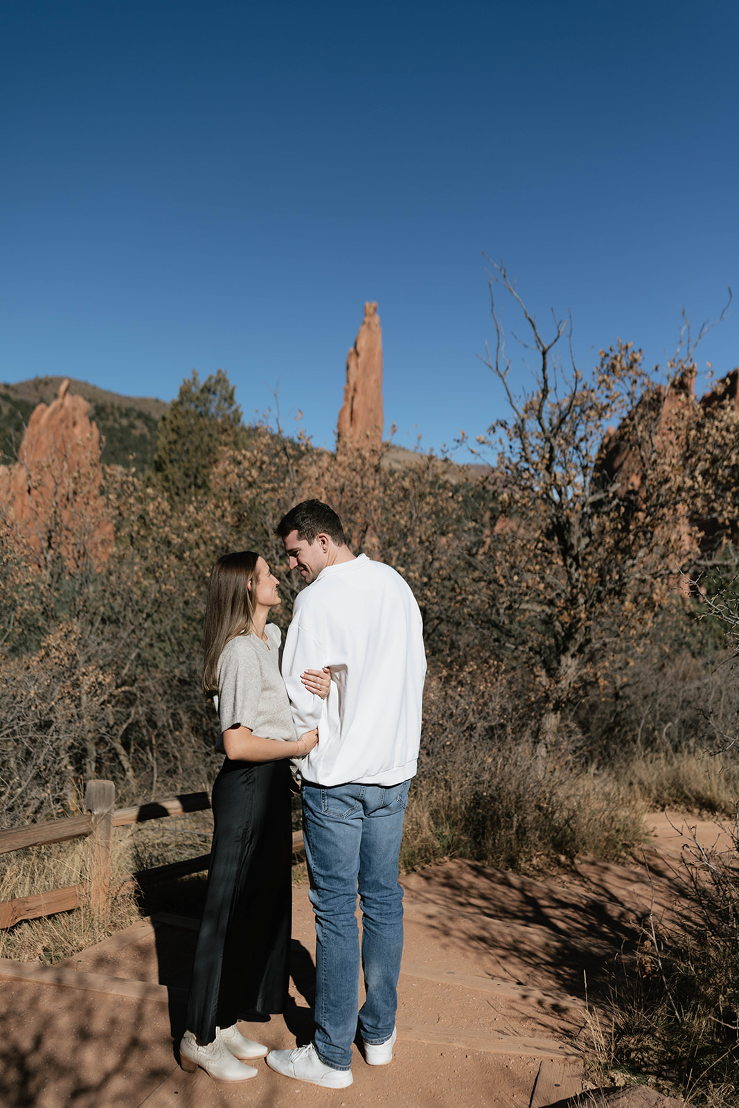 A couple smiling at each other on a trail overlook with jagged sandstone formations in the background at Garden of the Gods.