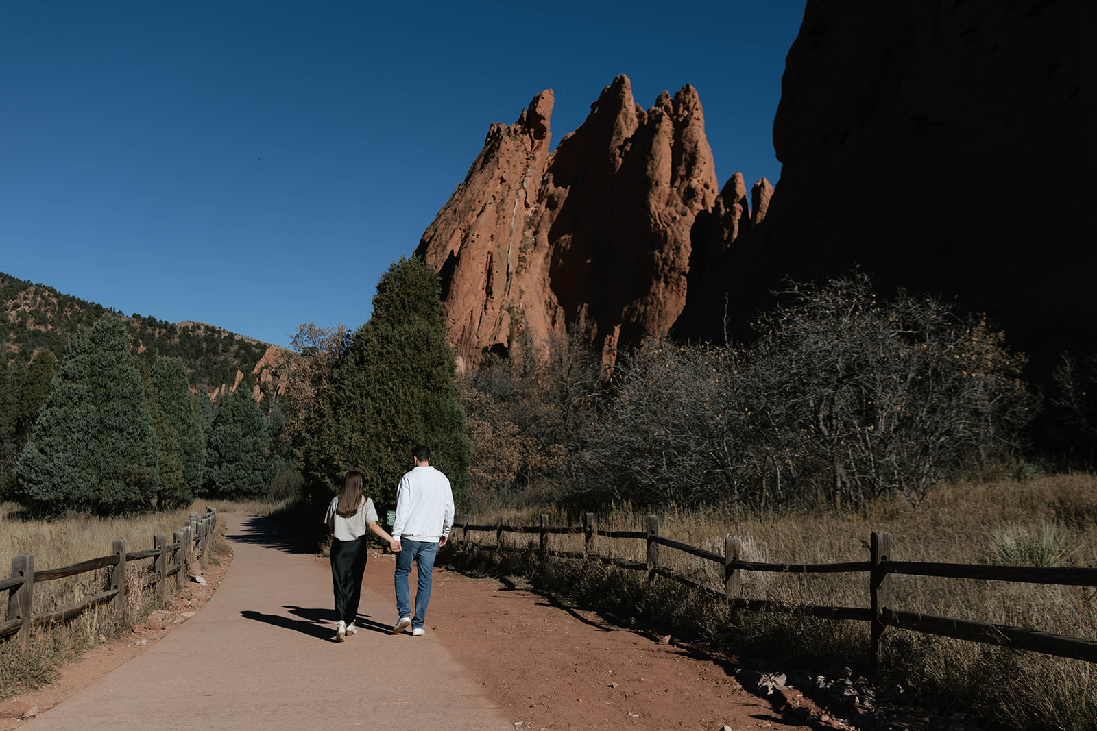 A couple strolling down a quiet path, hand-in-hand, with dramatic sandstone peaks rising in the distance at Garden of the Gods.