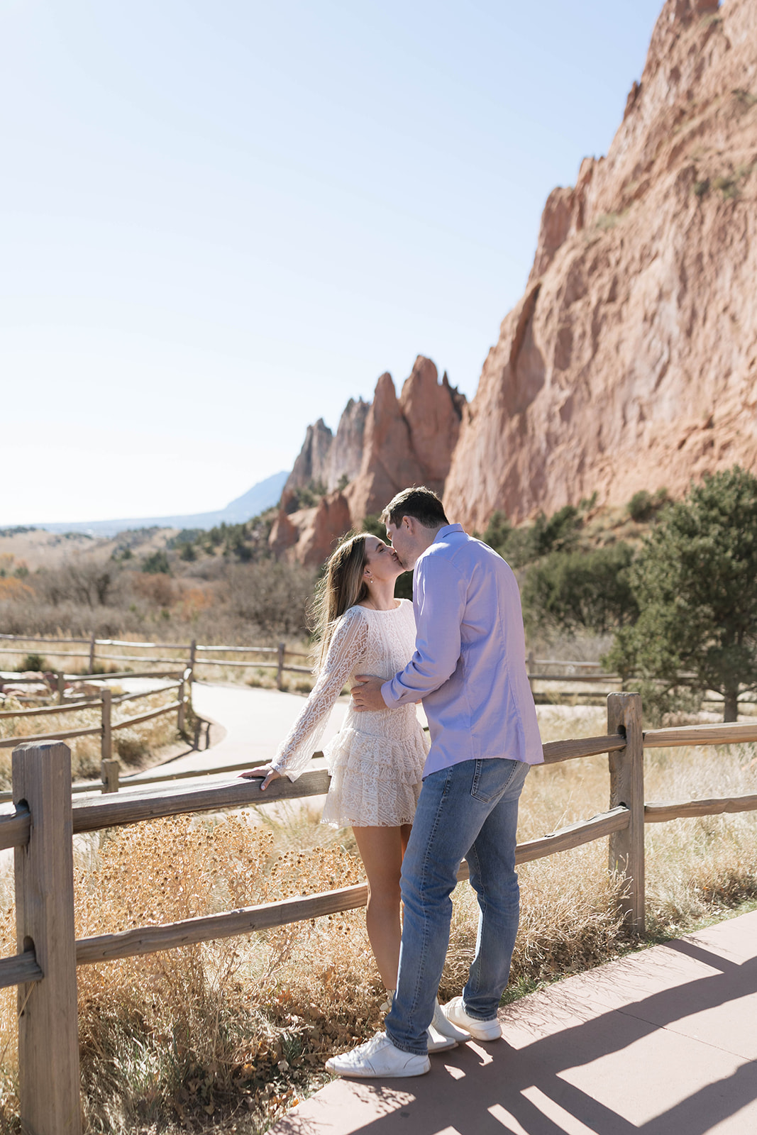 A couple standing beside a wooden fence, kissing each other with the towering sandstone cliffs of Garden of the Gods in the background.