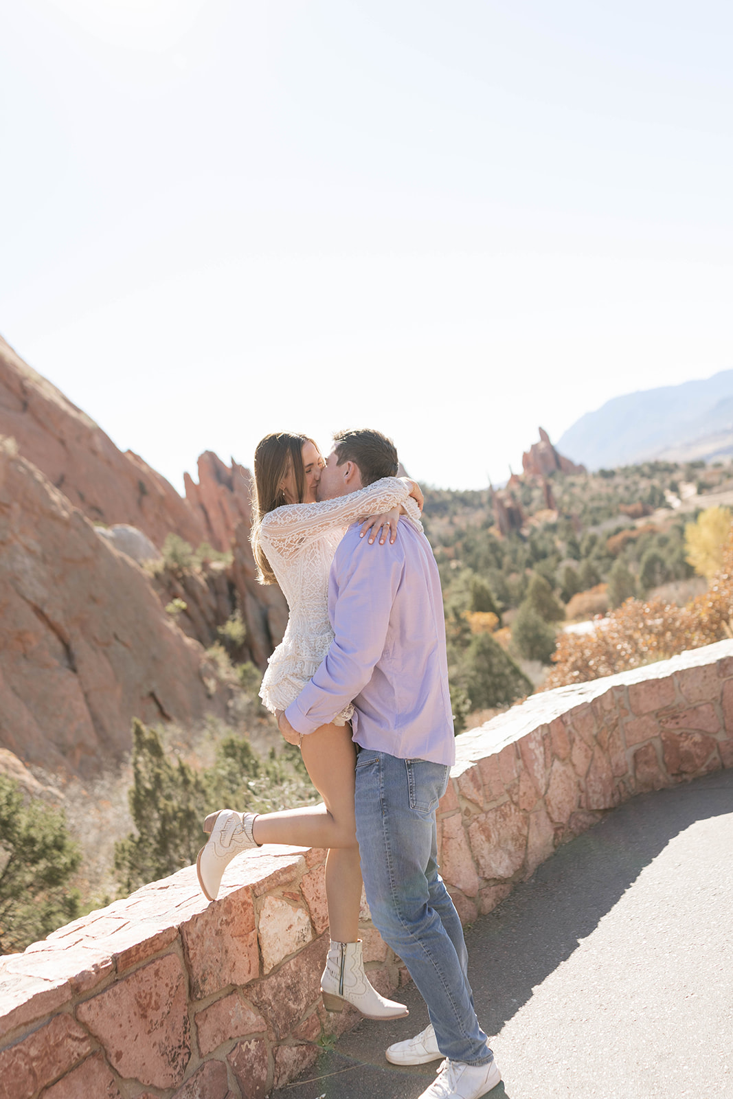 A joyful moment of a woman wrapped around her partner as they kiss beside a stone wall overlooking sweeping mountain and red-rock views.