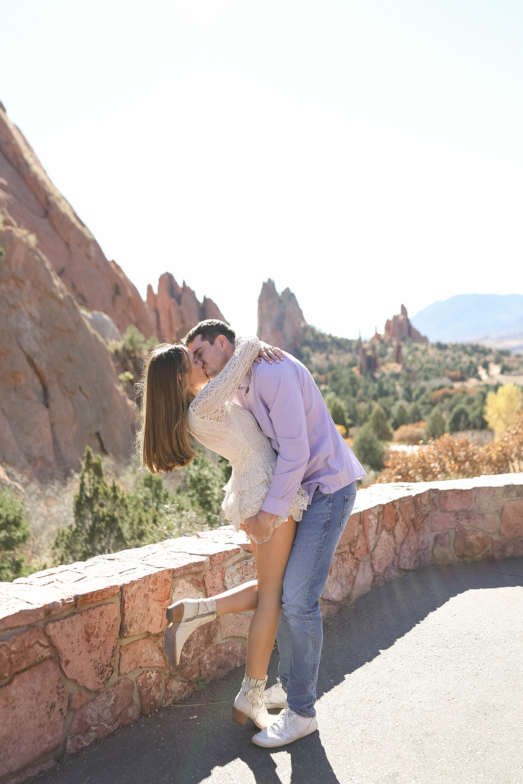 A couple sharing a romantic kiss as he lifts her beside a stone overlook with the red rock formations of Garden of the Gods stretching behind them.