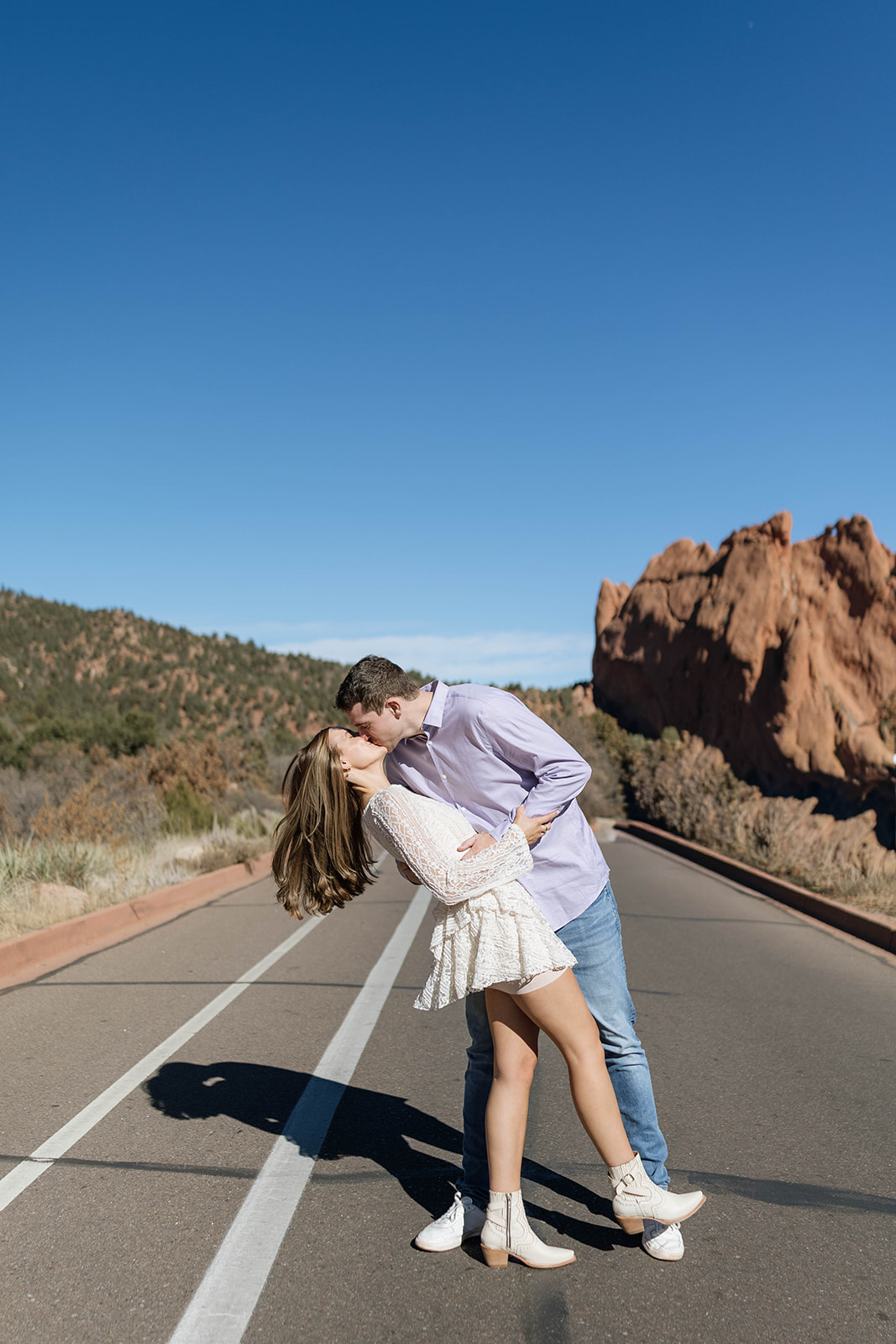 A couple sharing a dramatic dip-kiss in the middle of an empty road, the red rock formations of Garden of the Gods rising behind them under a clear blue sky.