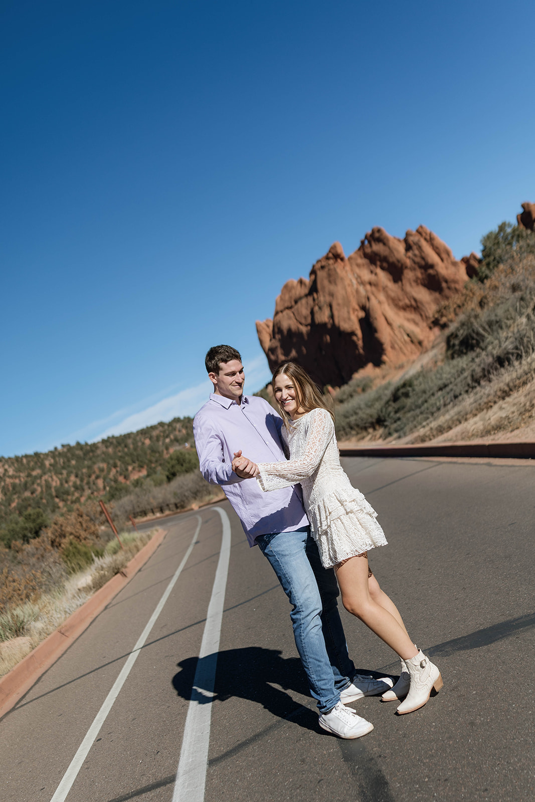 A couple smile while holding hands on a sunny road lined with towering sandstone at Garden of the Gods.