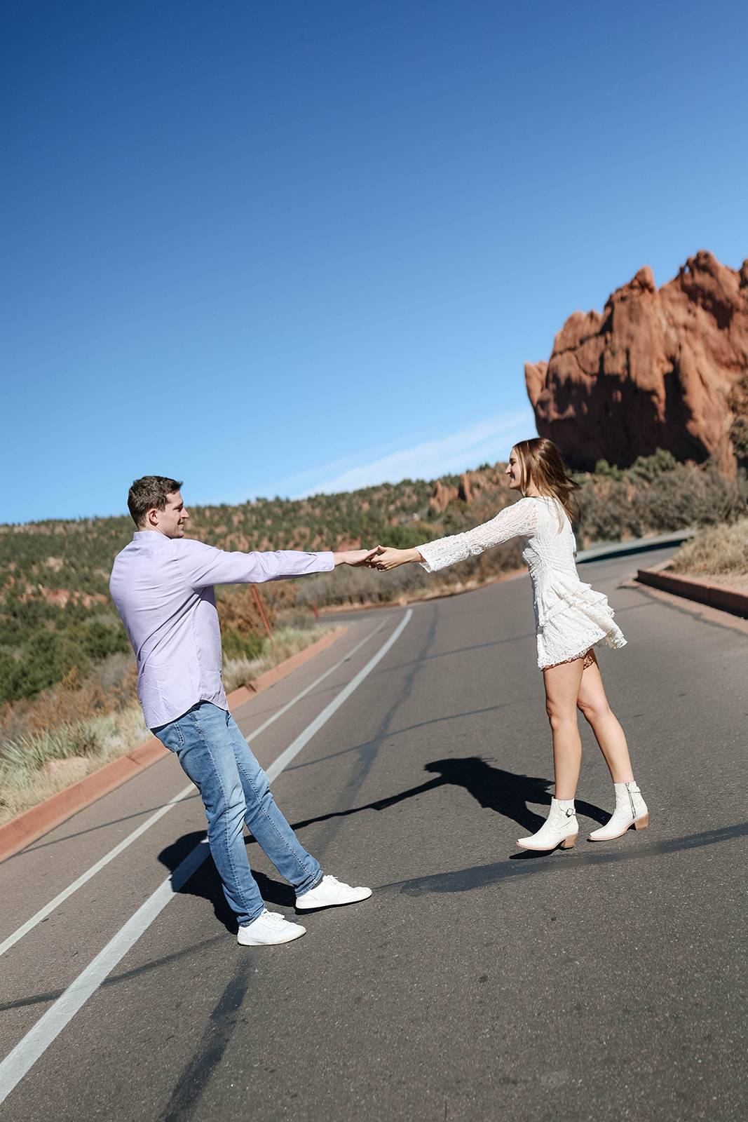 A playful moment of a couple spinning each other in the middle of a scenic roadway with Garden of the Gods peaks in the distance.