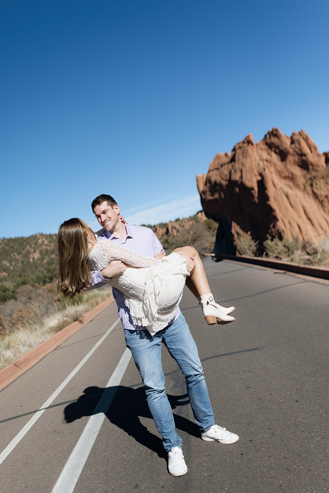 A man carrying his partner in his arms while they smile at each other on a sunny road lined with towering sandstone at Garden of the Gods.