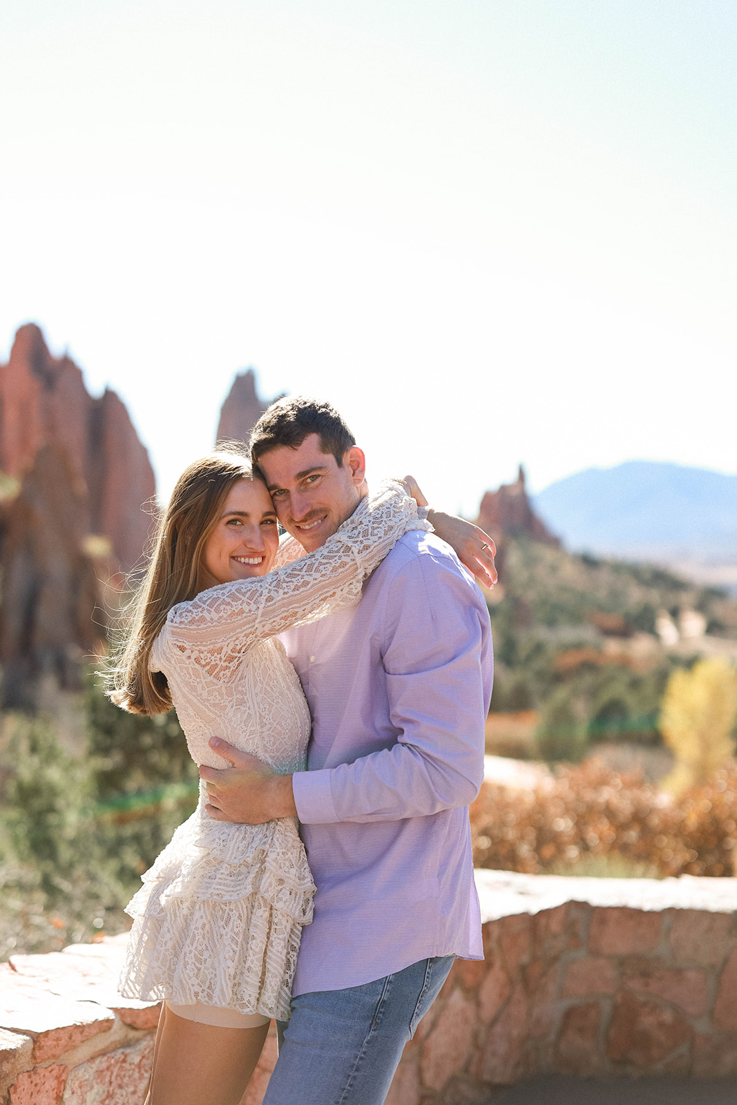 A couple hugging and smiling at the camera with sweeping rock formations of Garden of the Gods and mountain views behind them.