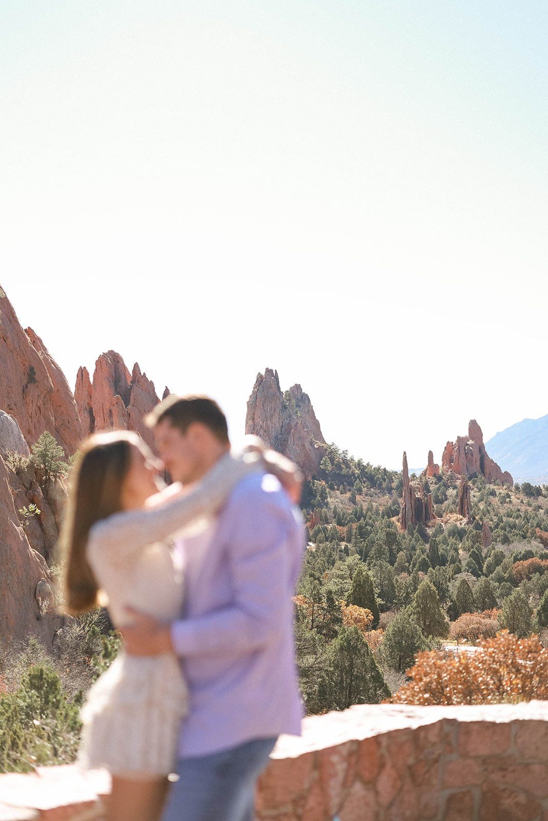 A blurred, intimate close-up of a couple holding each other with dramatic red rock spires stretching across the valley behind them.