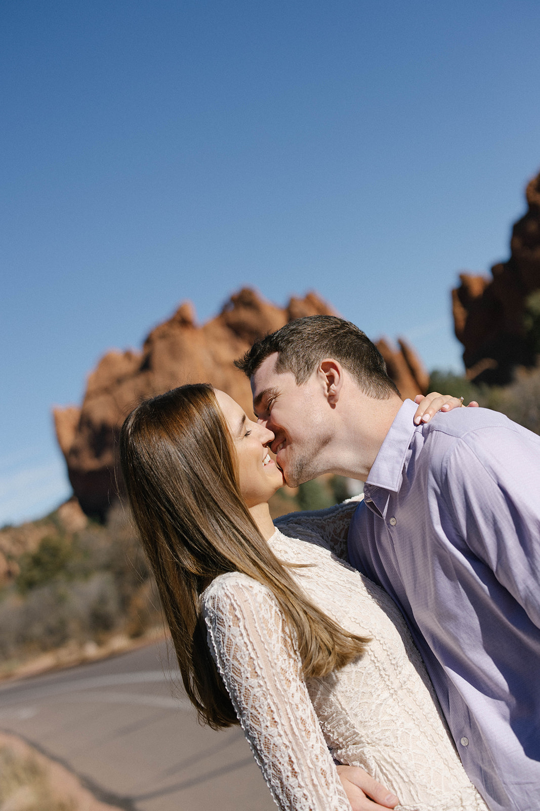 A close-up of a couple leaning in and kissing, smiling with red rock cliffs behind them.