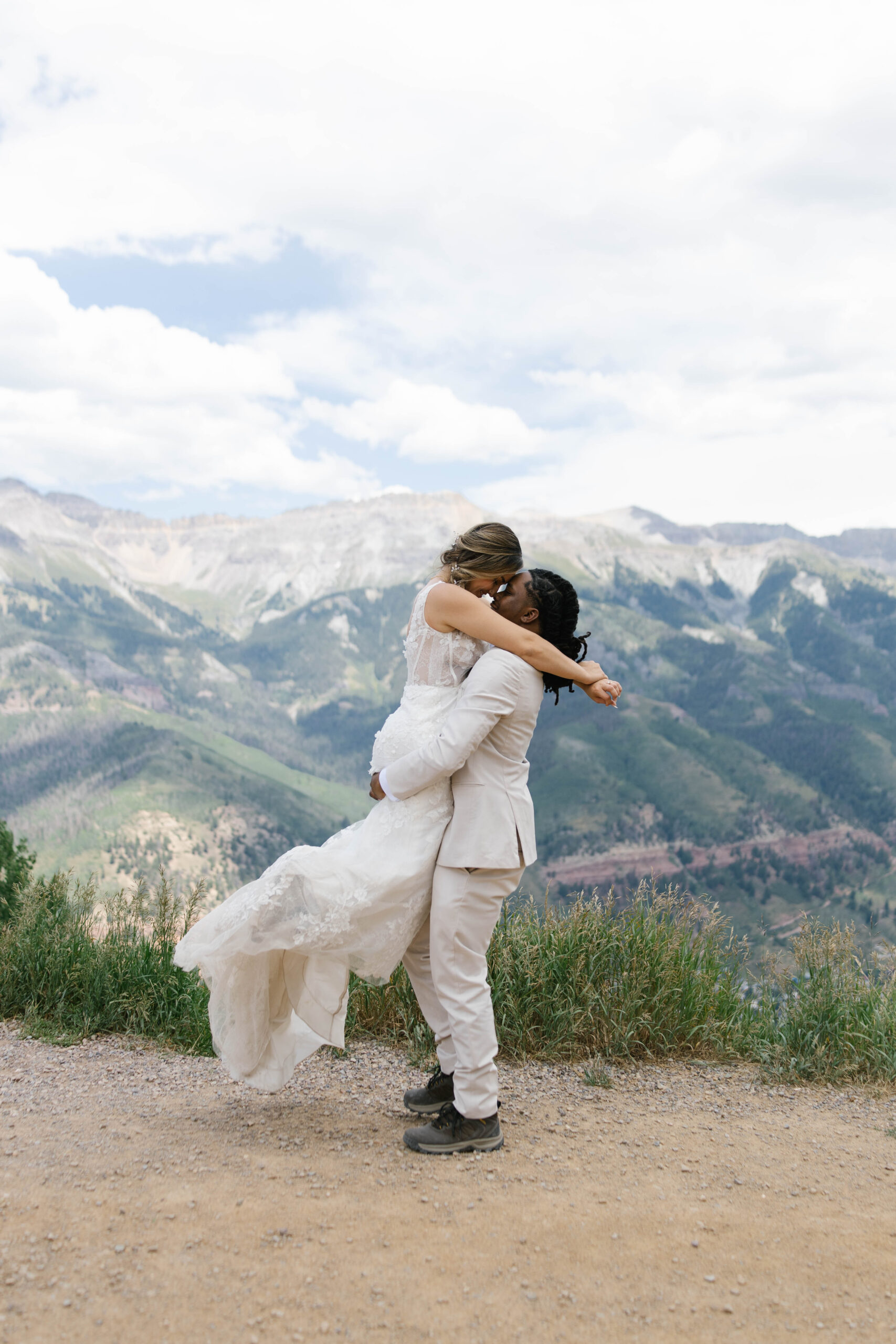 A groom picks up his bride beneath ski trail signs with expansive mountain ridges unfolding behind them.