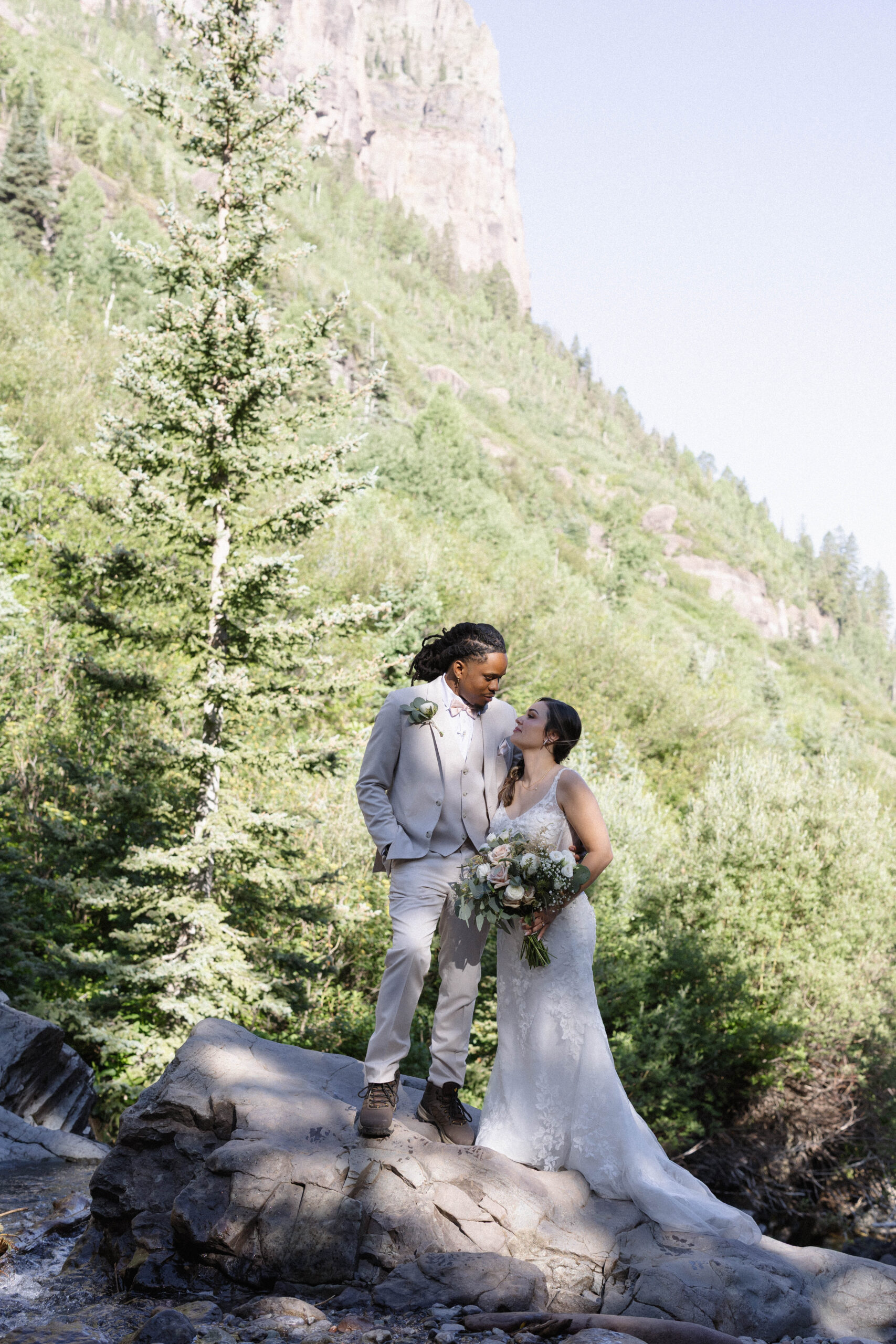 A couple standing together on a large rock surrounded by tall evergreens and rugged cliffs, sharing a quiet, intimate moment.